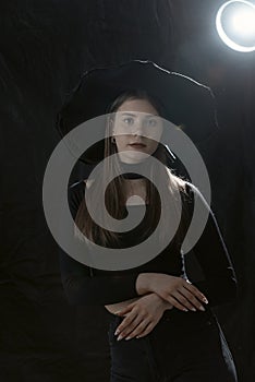 Portrait of young lady in black clothes and hat with wide brims. Black background. Young girl in Gothic style. Vertical frame