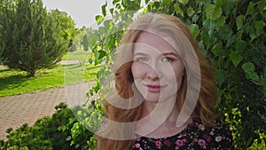 Portrait of young lady with long red hair and freckles posing happy state of mind looking at camera in park in summer