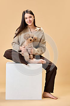 Portrait of young lady sitting on white cube, stage with her lovely cute, groomed Poodle against beige background