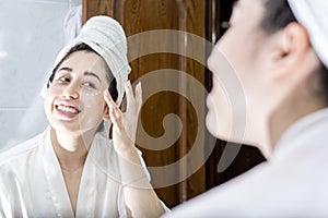 Portrait of a young Latin woman in a bathrobe and a towel on her head looking in the mirror while applying cream to her face