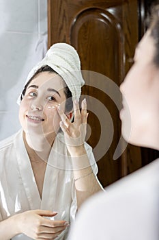 Portrait of a young Latin woman in a bathrobe and a towel on her head looking in the mirror while applying cream to her face