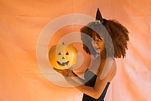 Portrait of young latina woman dressed as a witch holding a pumpkin with her hands on pink background for halloween