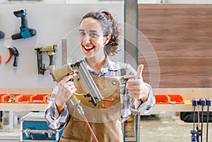 Portrait young latino carpenter holding nailer looking at camera smiling in furniture carpentry workshop