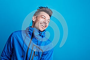 Portrait of a young laughing man in a studio with anorak on a blue background.