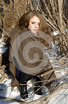 Portrait of young lonely girl sitting in winter snowy forest. Unhappy freezing woman.