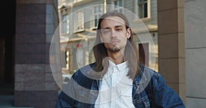 Portrait of young long haired man with earrings turning head and looking to camera. Close up view of bearded young man