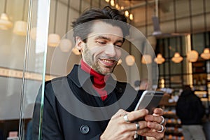 Portrait of young lovely brown haired unshaved male looking happily on screen of his phone and smiling cheerfully while standing