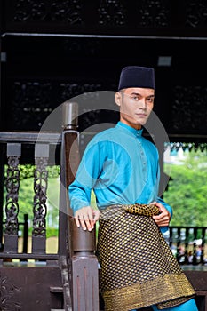 A portrait of young malay man in his traditional teal cloth, samping songket and songkok at traditional wooden house photo