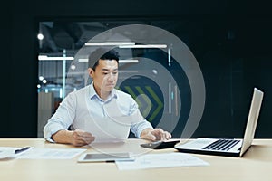 Portrait of a young male Asian accountant. He sits at the desk in the office, works with documents, accounts on a