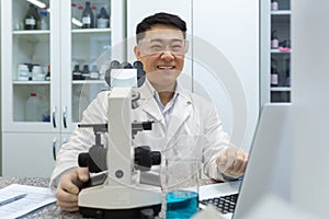 Portrait of young male Asian student, intern studying and working in laboratory with microscope and laptop. Sitting at