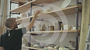 Portrait of young male ceramist, who is choosing mug to decorate it in his studio.