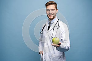 Portrait Of Young Male Doctor Holding Green Apple. Isolated On Light Blue.