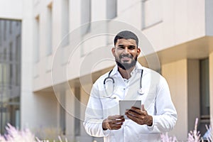 Portrait of a young male doctor, nurse, standing outside the hospital, holding a tablet and looking into the camera