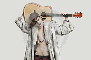 Portrait of young male guitarist in fur coat holding guitar over shoulder against gray background