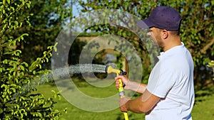 Portrait of young male holding hose and watering plant in garden with lake. Handsome man spraying water on green pine