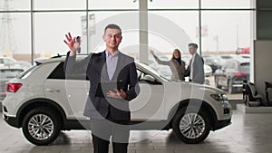 portrait of young male manager with keys in his hands backdrop of happy buyers of new car in showroom, smiling and