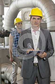 Portrait of young male manager writing on clipboard with manual worker in background at industry