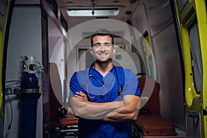 A portrait of a young male paramedic standing in front of an ambulance car with his hands folded