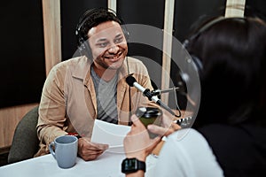 Portrait of young male radio host going live on air, talking with female guest, holding a script paper while sitting in