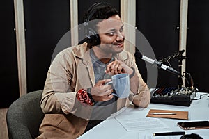 Portrait of young male radio host going live on air, talking with guest, holding a cup of coffee or tea while sitting in