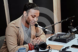 Portrait of young male radio host going live on air, talking in microphone, reading a script from paper while sitting in