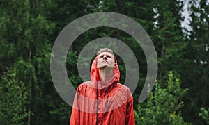 Portrait of a young male tourist in a red raincoat stands in the rain in the mountains against a background of trees, and looks up