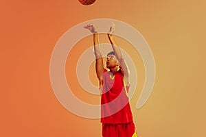 Portrait of young man, basketball player throwing ball into basket isolated over orange studio background in neon light