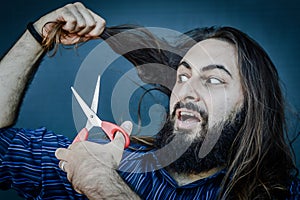 Portrait of a young man with a beard and long hair who, with a scissor in his hand, wants to change his look
