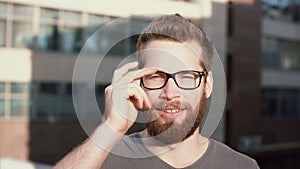 Portrait of young man with beard standing in front of sun and blinking. Attractive businessman takes off the glasses.