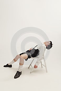 Portrait of young man in black and white outfit sitting on chair, posing  over grey studio background. Looks