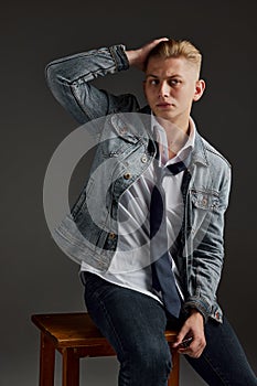 Portrait of young man with blonde hair posing in jeans jacket  over grey background. Casual style. Brutal look
