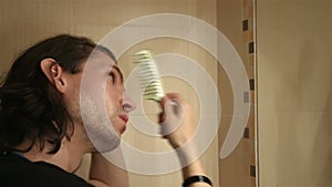 Portrait of young man brushing long layered dark brown hair in bathroom standing in front of mirror, side view, face profile