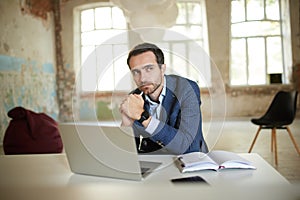 Portrait of young man, businessman sitting at the table with laptop, thinking and making notes. Empty building