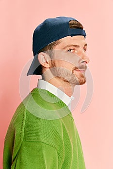 Portrait of young man in cap posing in green sweater and shirt isolated over pink background. Side view