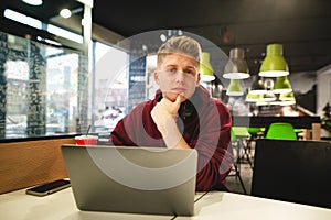 Portrait of a young man in casual clothing sitting with a laptop in a fast food cafe, looking at the camera