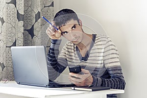 Portrait of young man In casual wear on the job working on laptop, using  smartphone and looking at with thoughtful face