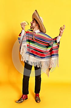 Portrait of young man in colorful clothes, poncho and sombrero posing, eating taco against yellow studio background