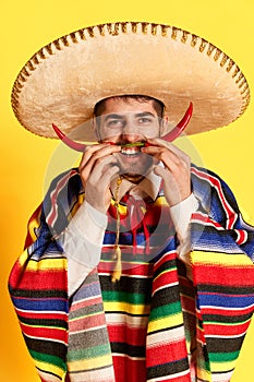 Portrait of young man in colorful festive clothes, poncho and sombrero posing with chilli pepper like moustaches against