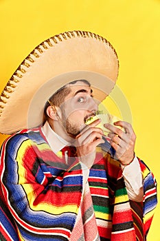 Portrait of young man in colorful festive clothes, poncho and sombrero posing, eating taco against yellow studio