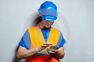 Portrait of young man construction worker engineer, typing on smartphone, wearing safety equipment on grey textured background.