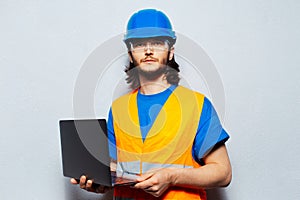 Portrait of young man construction worker engineer wearing safety equipment, holding laptop on background of grey textured wall.