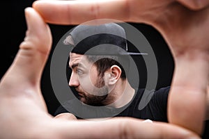 Portrait of young man with dark hair in shirt and hat on black background. Man`s hands making frame