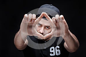 Portrait of young man with dark hair in shirt and hat on black background. Man`s hands making frame