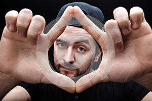 Portrait of young man with dark hair in shirt and hat on black background. Man`s hands making frame