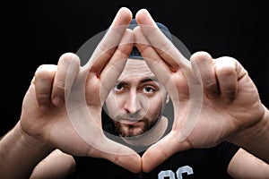 Portrait of young man with dark hair in shirt and hat on black background. Man`s hands making frame