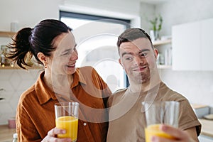 Portrait of young man with Down syndrome with his mother at home, toasting with juice. Morning routine for man with Down