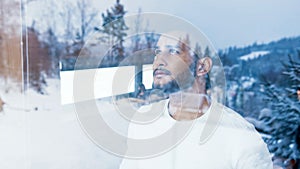 Portrait of young man drinking hot beverage wile looking through the window at first snow. Christmas celebration