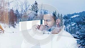 Portrait of young man drinking hot beverage wile looking through the window at first snow. Christmas celebration