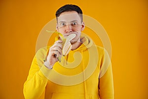 Portrait of young man eating banana on yellow background. Funny guy with bulging eyes in yellow hoodie shoved fruit in