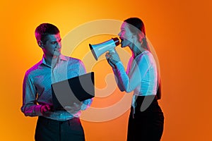 Portrait of young man, employee with laptop and woman shouting in megaphone isolated over orange background in neon